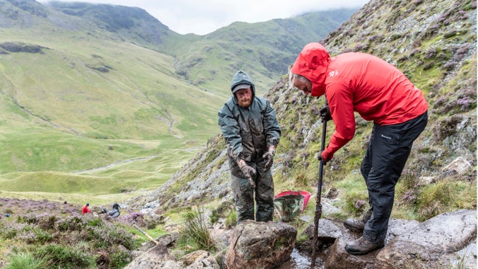 Two men dressed in wet waterproofs, one blue, one red, work to lever rocks into places to repair a footpath in open countryside, an empty valley stretching behind them under grey rain clouds.
